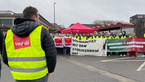 A man is wearing a yellow Ver.di vest.  In the background several strikers with posters.  © NDR Photo: Christian Nagel