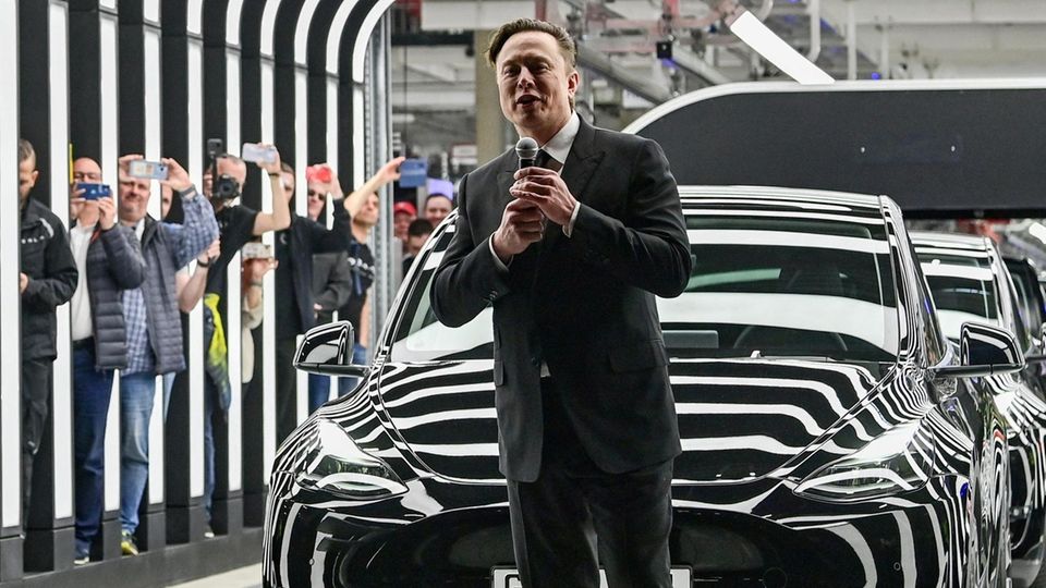 Elon Musk with a microphone in front of a black Tesla at an event