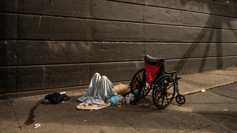 A man lies on the street behind a wheelchair, covered only by a hospital blanket