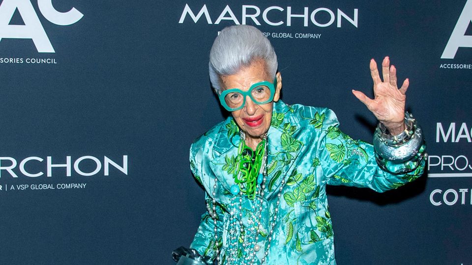 Iris Apfel waving and in a floral suit