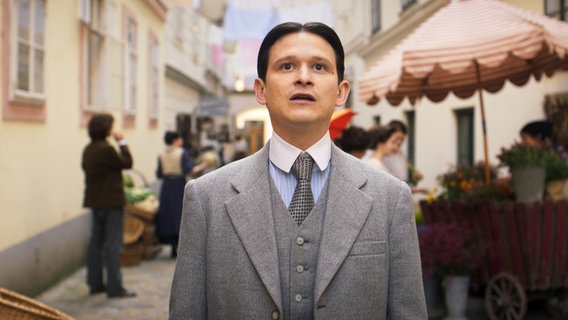A man with dark hair and a middle part in a gray suit at a flower market looks up in amazement - Franz Kafka (Joel Basman) in the series "Kafka" © NDR/Superfilm 