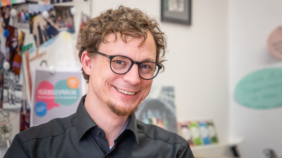 Happiness researcher and psychologist Tobias Rahm, a man in a black shirt, glasses and curly hair, smiles into the camera