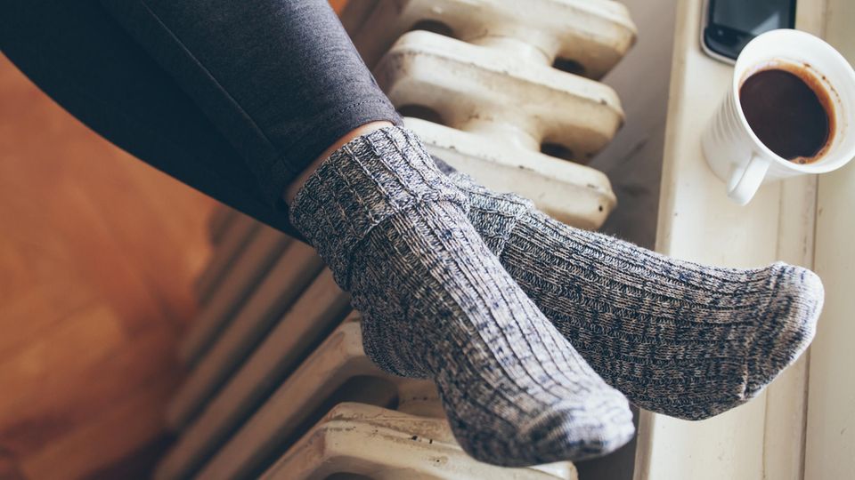 A woman's feet lie on a radiator in wool socks.  There is a coffee mug and a smartphone on the windowsill